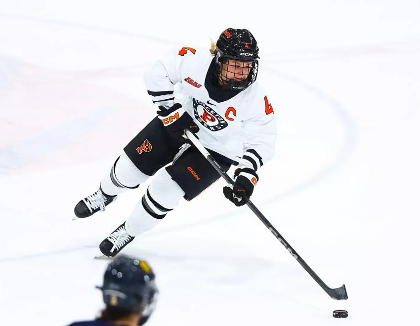 Mia Coene in a white Princeton Ice Hockey uniform skating with control of the puck against a nearly clear field of ice.