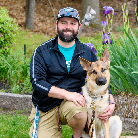 Jeffrey, on one knee, looks at te camera smiling with a german shephard sitting on his left; head slightly cocked to the side looking inqusisitively at the camera